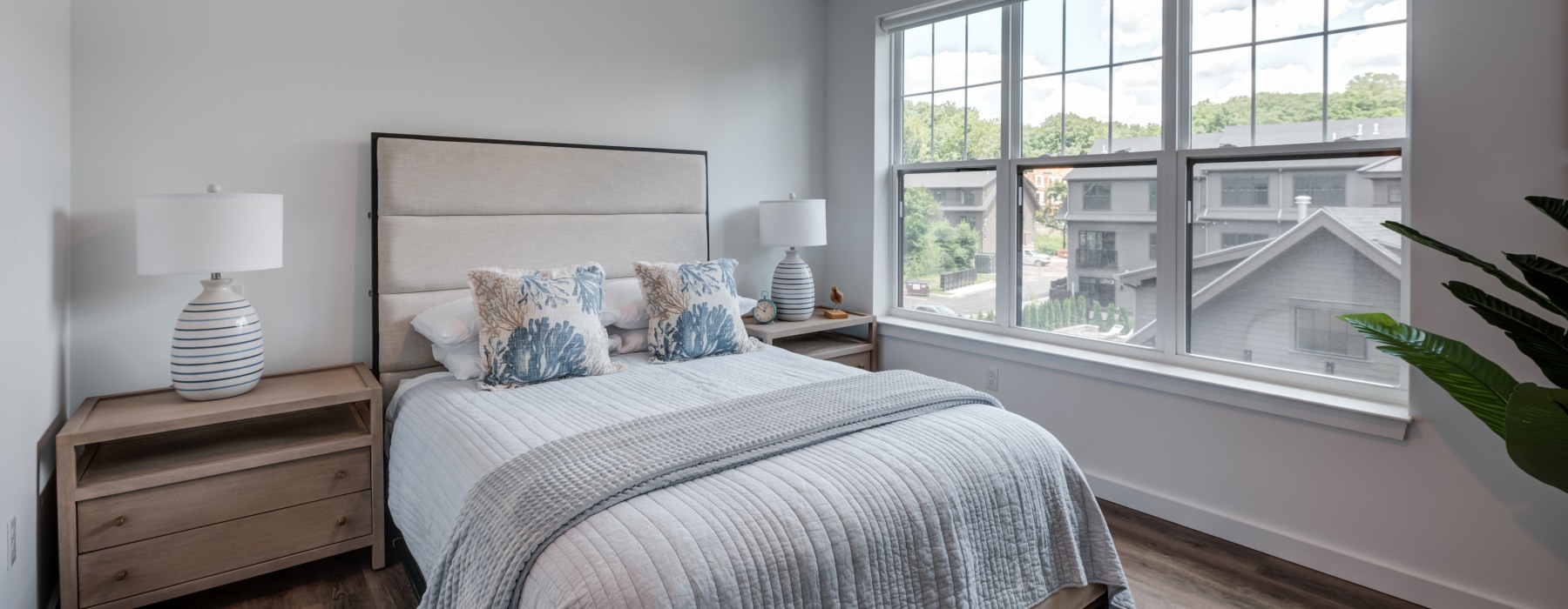 Bright, modern model apartment bedroom with cozy bedding, neutral decor, and a large window at Brookside Wilton in CT